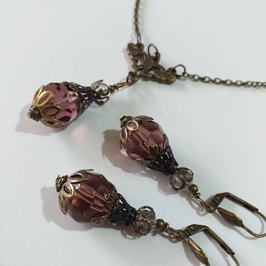 Op de afbeelding: Een set met paarse glazen en bronzen oorbellen en een bijpassende ketting. De oorbellen hebben een traanvorm met een bloemmotief en de ketting heeft een Y-vormig ontwerp met een bloemvormige hanger.
