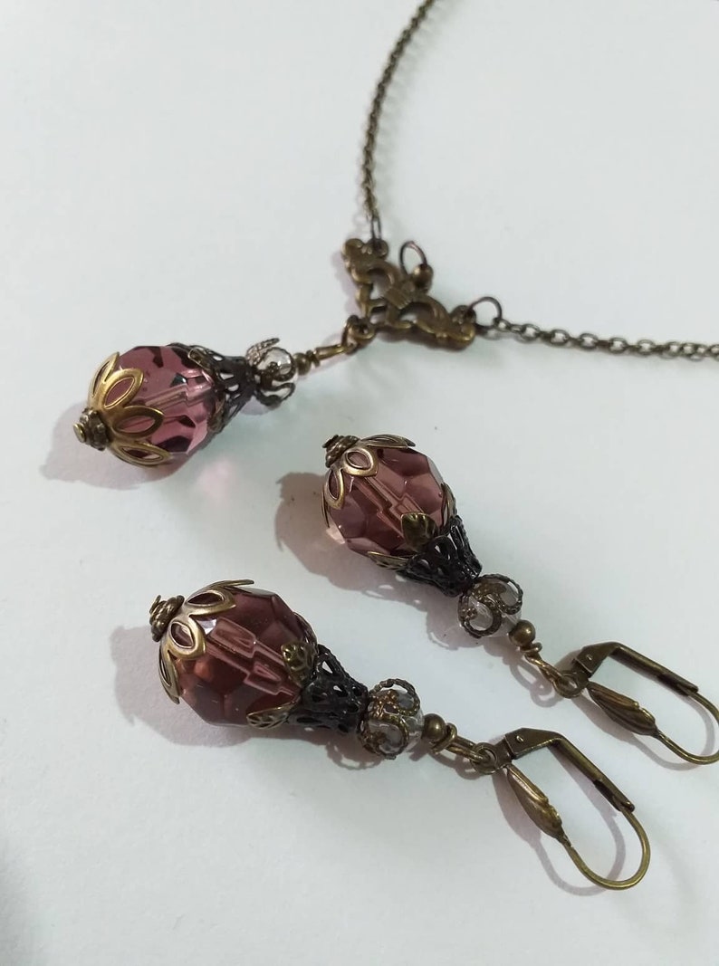 Op de afbeelding: Een set met paarse glazen en bronzen oorbellen en een bijpassende ketting. De oorbellen hebben een traanvorm met een bloemmotief en de ketting heeft een Y-vormig ontwerp met een bloemvormige hanger.
