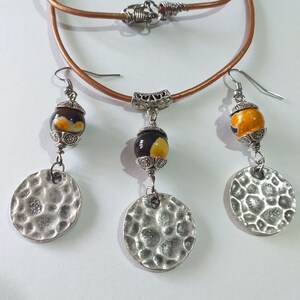 May include: A jewelry set featuring a brown leather necklace with a silver clasp, and matching earrings. The earrings and necklace pendant have a decorative bead with yellow and brown accents, and a hammered silver disc. The set is displayed on a white background.