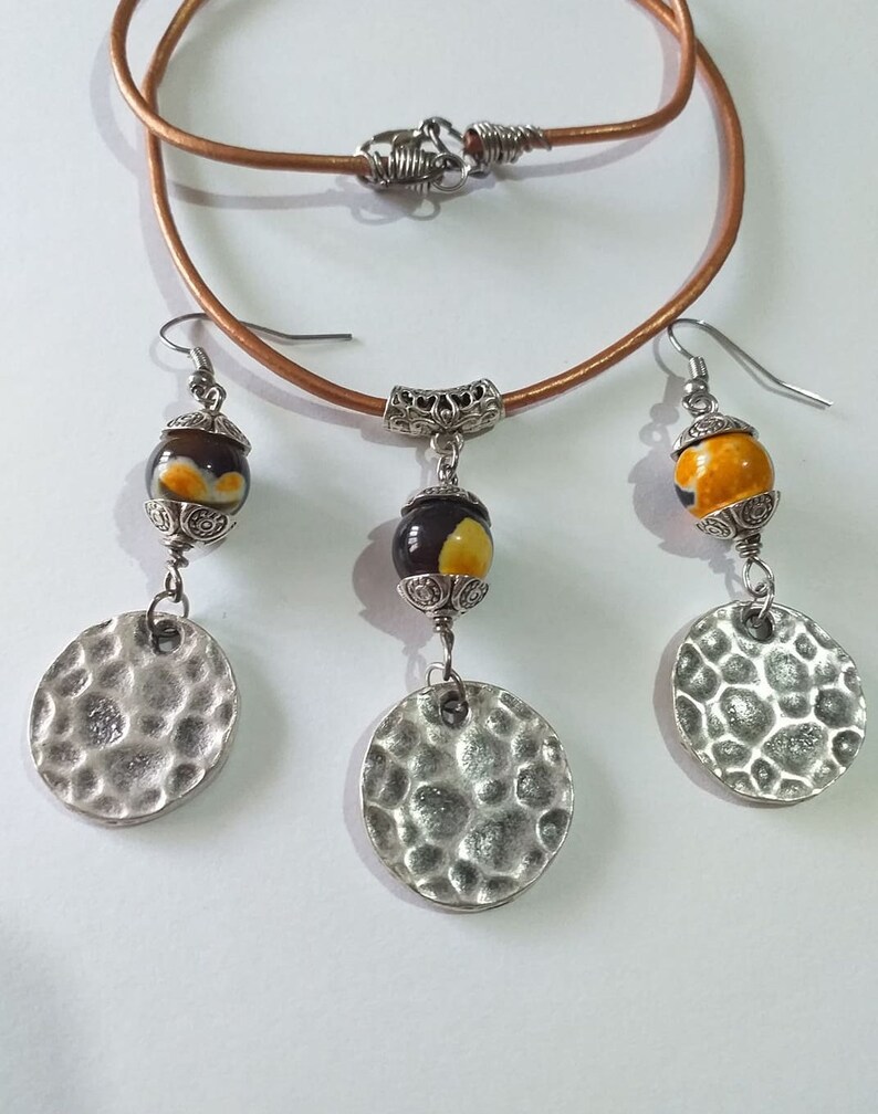 May include: A jewelry set featuring a brown leather necklace with a silver clasp, and matching earrings. The earrings and necklace pendant have a decorative bead with yellow and brown accents, and a hammered silver disc. The set is displayed on a white background.