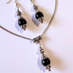 May include: A silver necklace and matching earrings set. The necklace features a pendant with a large, round, black bead. The earrings have a similar design, with a black bead and silver accents. The jewelry set is displayed on a white background.