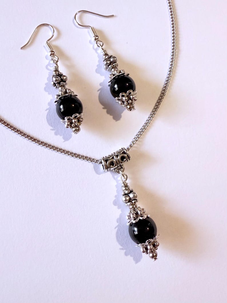May include: A silver necklace and matching earrings set. The necklace features a pendant with a large, round, black bead. The earrings have a similar design, with a black bead and silver accents. The jewelry set is displayed on a white background.
