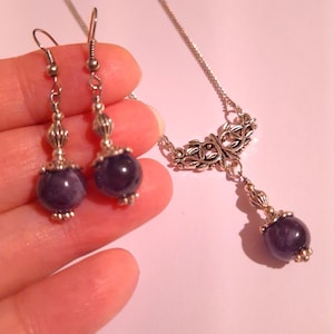May include: A silver necklace and earrings set with a purple stone pendant and matching earrings. The necklace features a delicate, ornate design with a dangling stone.