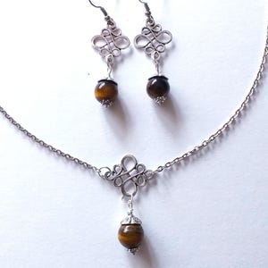 May include: A silver necklace and earrings set with a brown tiger's eye stone pendant and matching earrings. The necklace features a silver chain and a decorative silver pendant with a knotted design. The earrings have a similar knotted design with a brown tiger's eye stone dangling from each.