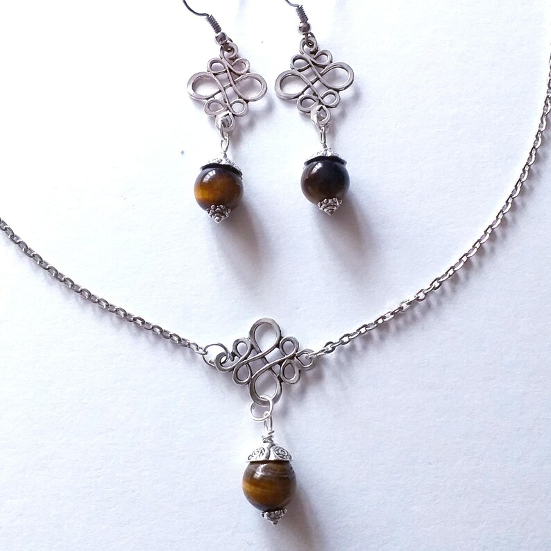 May include: A silver necklace and earrings set with a brown tiger's eye stone pendant and matching earrings. The necklace features a silver chain and a decorative silver pendant with a knotted design. The earrings have a similar knotted design with a brown tiger's eye stone dangling from each.