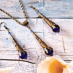 May include: A set of blue glass and gold tone earrings and necklace. The earrings and necklace feature a teardrop shaped pendant with intricate filigree details.