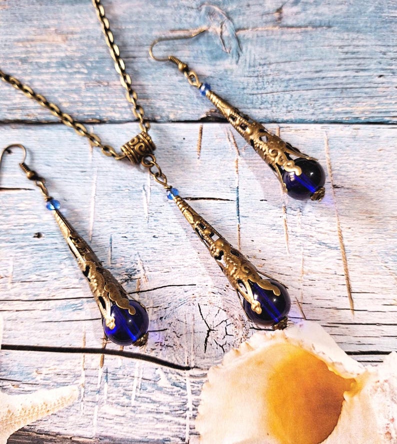 May include: A set of blue glass and gold tone earrings and necklace. The earrings and necklace feature a teardrop shaped pendant with intricate filigree details.