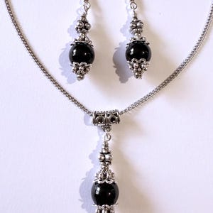 May include: A silver-toned necklace and earring set. The necklace features a chain with a pendant of a black bead with silver accents. The earrings have a similar design, with black beads and silver decorative elements. The jewelry set is displayed against a white background.
