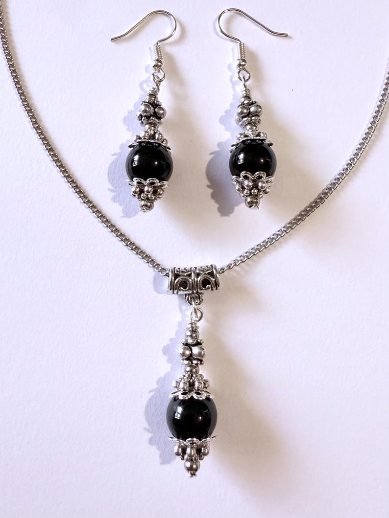 May include: A silver-toned necklace and earring set. The necklace features a chain with a pendant of a black bead with silver accents. The earrings have a similar design, with black beads and silver decorative elements. The jewelry set is displayed against a white background.