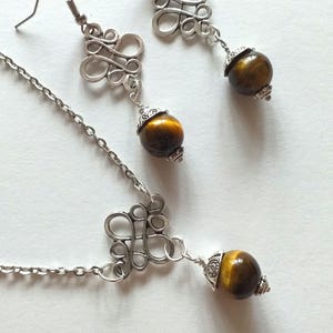May include: A silver chain necklace with a silver pendant featuring a decorative knot design. The pendant is attached to a round tiger's eye gemstone. Matching earrings with a similar design and tiger's eye gemstones are also included.