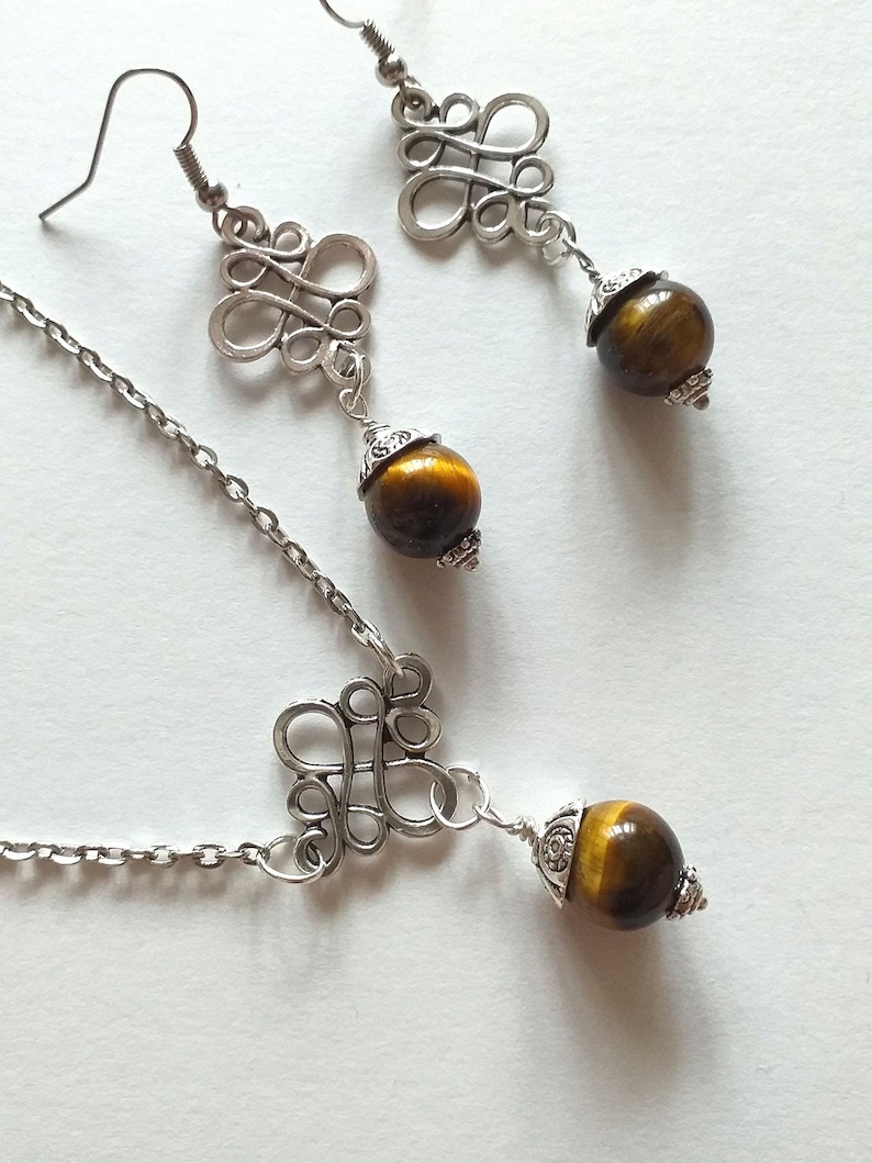May include: A silver chain necklace with a silver pendant featuring a decorative knot design. The pendant is attached to a round tiger's eye gemstone. Matching earrings with a similar design and tiger's eye gemstones are also included.
