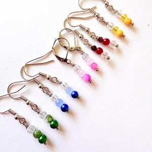 May include: A collection of six pairs of drop earrings featuring silver-toned metal hooks and lever backs. Each earring is adorned with a clear faceted bead followed by two round coloured beads in yellow, red, pink, blue, and green hues.