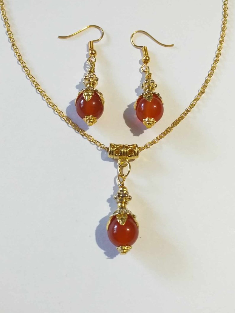 K&ouml;nnte beinhalten: Eine goldene Halskette mit einem roten Karneol-Anh&auml;nger und passenden Ohrringen. Anh&auml;nger und Ohrringe sind mit goldenen Akzenten verziert.