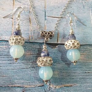 May include: A silver necklace and earrings set with blue and white gemstone beads. The necklace has a silver chain and a silver pendant with a blue and white gemstone bead. The earrings have silver hooks and blue and white gemstone beads.