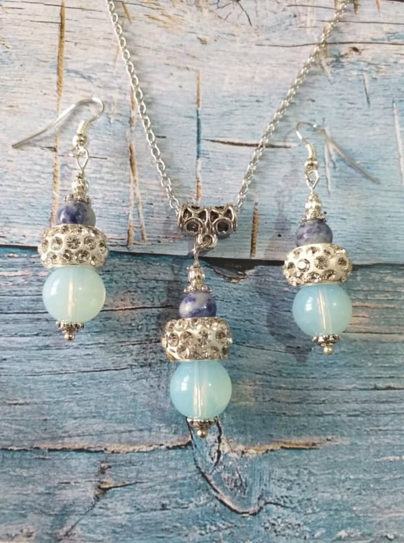 May include: A silver necklace and earrings set with blue and white gemstone beads. The necklace has a silver chain and a silver pendant with a blue and white gemstone bead. The earrings have silver hooks and blue and white gemstone beads.