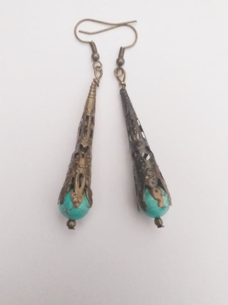 Puede incluir: Un par de pendientes colgantes con cuentas de color azul turquesa y metal con trabajo intrincado. El metal es de color dorado y tiene un dise&ntilde;o de filigrana.