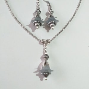 May include: A silver necklace and earrings set featuring grey glass beads and silver accents. The necklace has a delicate chain and a pendant with a teardrop-shaped bead. The earrings are drop earrings with a similar teardrop-shaped bead.