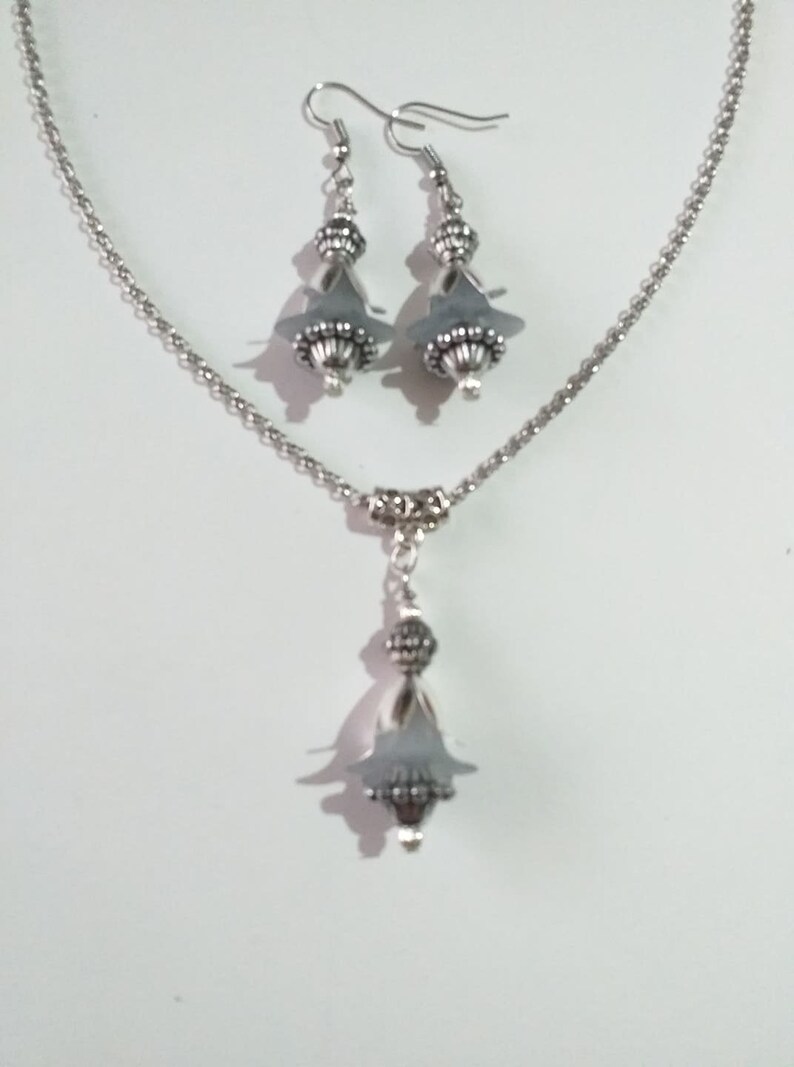 May include: A silver necklace and earrings set featuring grey glass beads and silver accents. The necklace has a delicate chain and a pendant with a teardrop-shaped bead. The earrings are drop earrings with a similar teardrop-shaped bead.