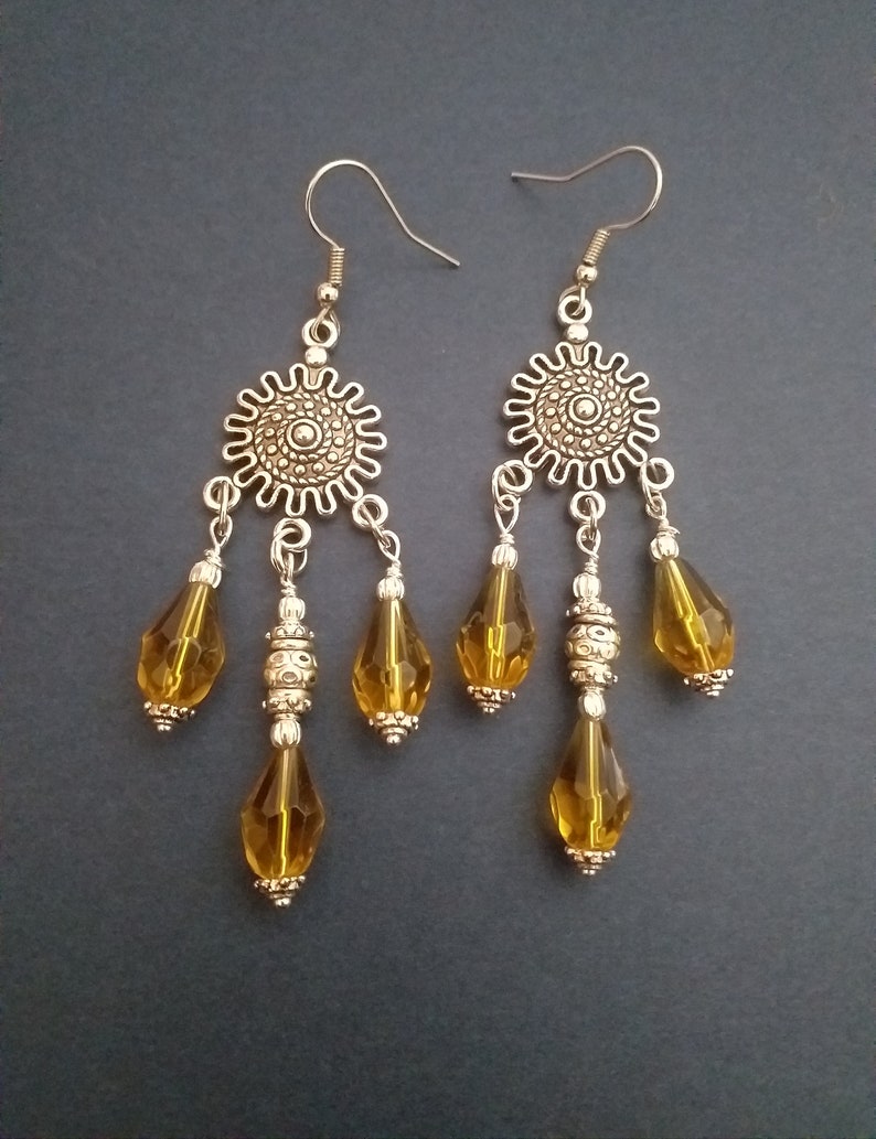 May include: A pair of dangle earrings with a silver tone finish. Each earring features a round, ornate charm with three teardrop-shaped amber glass beads hanging from it.