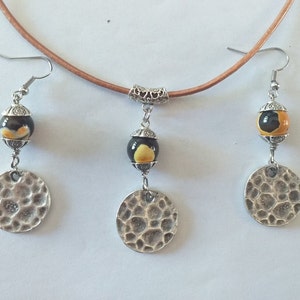 May include: A handcrafted jewelry set featuring a brown leather cord necklace with a pendant and matching earrings. The earrings and pendant have a black and yellow bead, and a hammered silver-tone disc. The set is displayed on a white background.