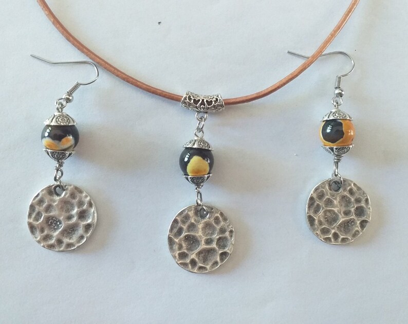May include: A handcrafted jewelry set featuring a brown leather cord necklace with a pendant and matching earrings. The earrings and pendant have a black and yellow bead, and a hammered silver-tone disc. The set is displayed on a white background.