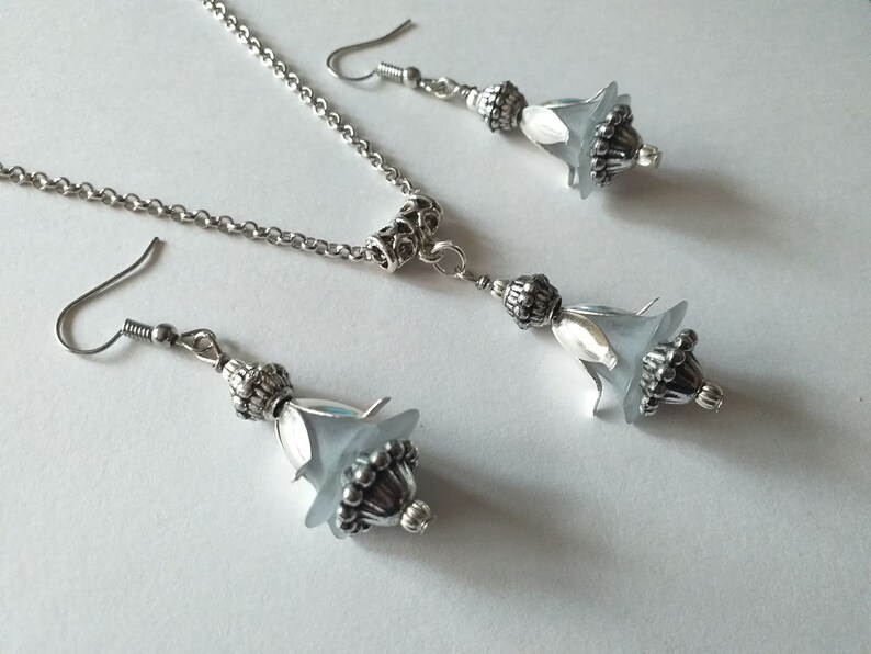 May include: A silver chain necklace and matching earrings with a frosted glass flower pendant. The flower pendant has a silver bead detail.
