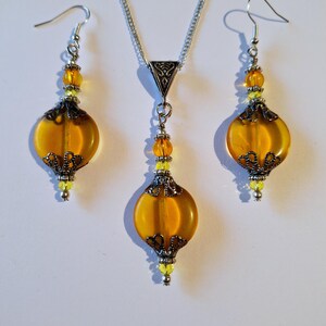 May include: A set of amber glass earrings and a necklace with silver chain and accents. The earrings and pendant feature a round, slightly flattened amber glass bead with a silver filigree design.