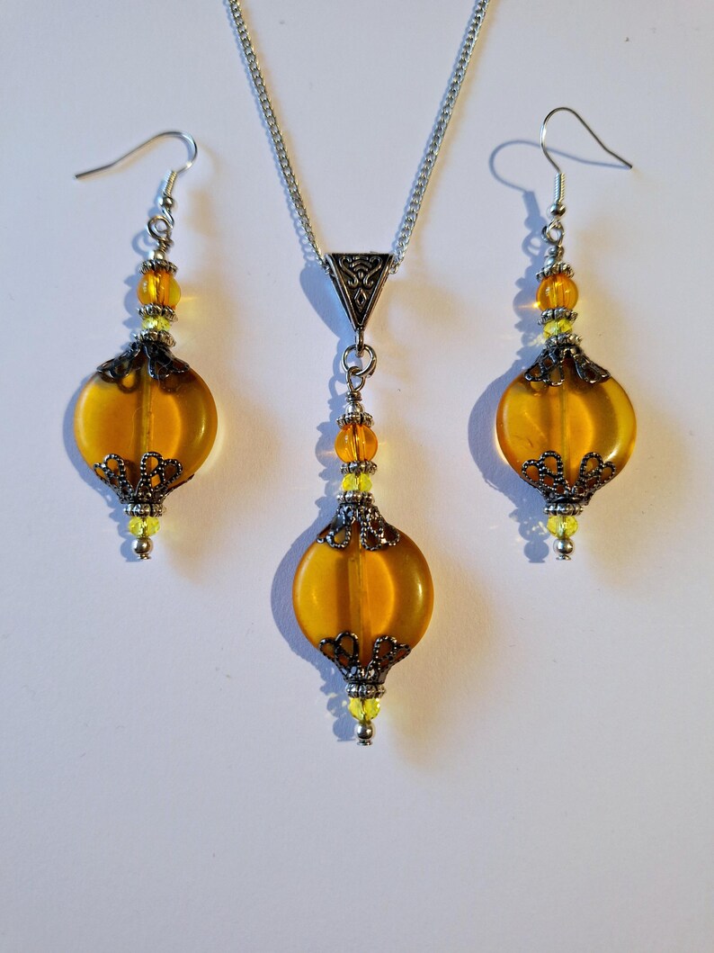 May include: A set of amber glass earrings and a necklace with silver chain and accents. The earrings and pendant feature a round, slightly flattened amber glass bead with a silver filigree design.