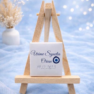 May include: A small wooden easel displays a white square with elegant blue script reading "Urime Synetia Orian" and the date "14.12.2023", along with an evil eye symbol. The background is a soft blue with bokeh lights.