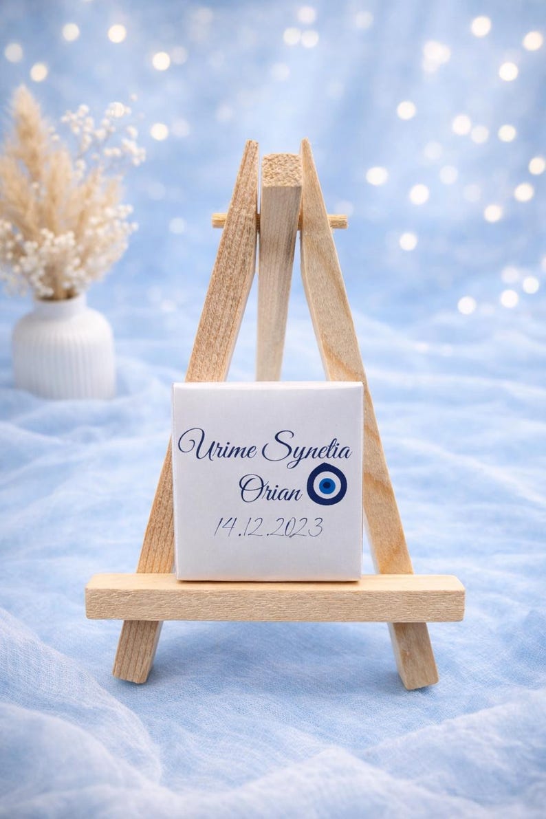 May include: A small wooden easel displays a white square with elegant blue script reading "Urime Synetia Orian" and the date "14.12.2023", along with an evil eye symbol. The background is a soft blue with bokeh lights.