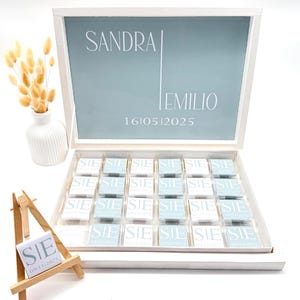 Può includere: Una scatola bianca con interno azzurro, personalizzata con i nomi "SANDRA" e "EMILIO" e la data "1610512025". La scatola contiene piccole caramelle incartate. Un piccolo cavalletto di legno mostra una caramella con le lettere "SIE" e la data "09112025".