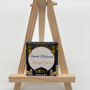 K&ouml;nnte beinhalten: Ein kleiner Holzstaffelei mit einer Karte in Gold- und Schwarzdesign. Die Karte tr&auml;gt den Text "Umrah Mubarak Zakariya Qassem" und zeigt eine Darstellung der Kaaba.