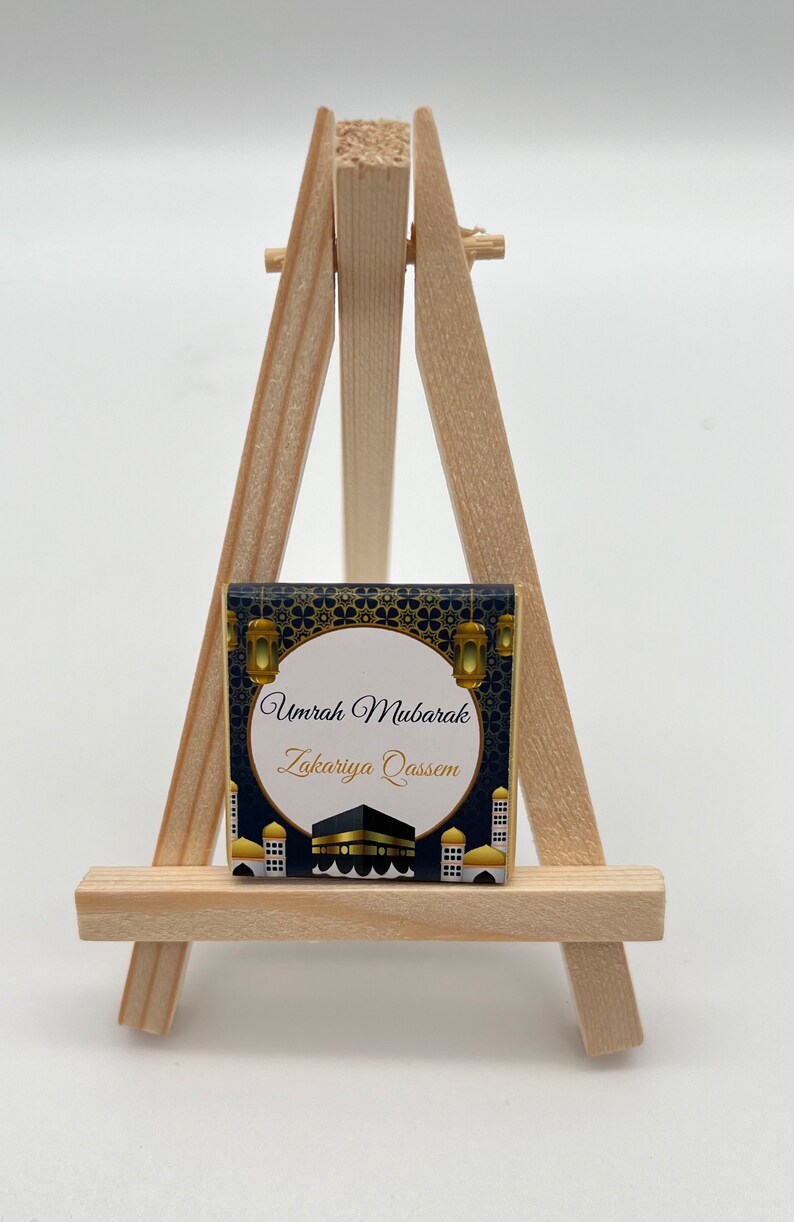 K&ouml;nnte beinhalten: Ein kleiner Holzstaffelei mit einer Karte in Gold- und Schwarzdesign. Die Karte tr&auml;gt den Text "Umrah Mubarak Zakariya Qassem" und zeigt eine Darstellung der Kaaba.