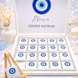 May include: A white wooden box filled with small, square chocolates, each adorned with a blue and white evil eye design. The box lid features a larger evil eye and the text "Elias'in Sünnet Hatırası". A small easel displays a similar chocolate.