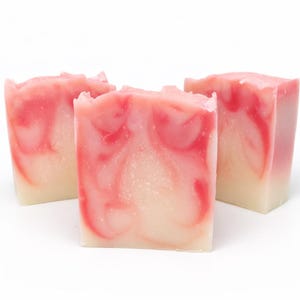 May include: Three handmade soap bars with a layered design. The soap features a creamy white base with swirling patterns of pink and red. The bars have a rectangular shape and a textured, uneven top.