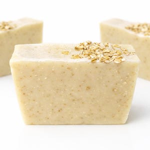 May include: Three rectangular bars of handmade soap are displayed against a white background. The soap is a light beige color, speckled with small particles, and topped with a scattering of rolled oats. The soap has a rustic, natural appearance.