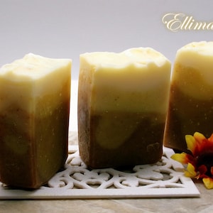 May include: Three rectangular bars of soap with a layered color scheme of brown, yellow, and white. The soap bars are arranged on a white decorative surface, with sunflowers on either side. The text "Ellimas Palm" is in the upper right corner.