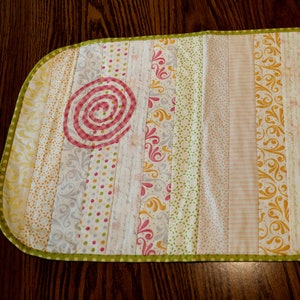 Puede incluir: Un camino de mesa rectangular largo con un borde verde. El camino de mesa est&aacute; hecho de tela con un dise&ntilde;o de patchwork de diferentes patrones florales en tonos de rosa, amarillo y verde. Hay dos dise&ntilde;os grandes en espiral en el centro del camino de mesa, uno en rosa y otro en naranja.