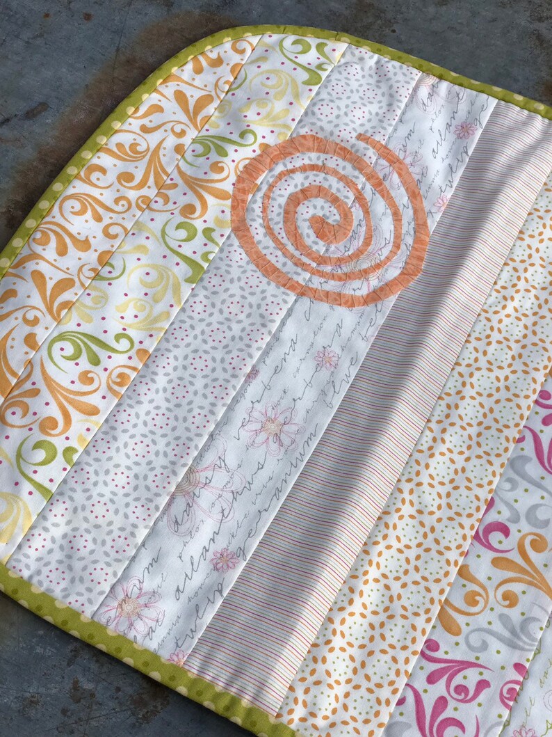 Puede incluir: Un posavasos acolchado con un borde verde y un dise&ntilde;o en espiral en el centro. El posavasos est&aacute; hecho de varias telas con patrones florales y geom&eacute;tricos en tonos de blanco, amarillo, rosa y verde.