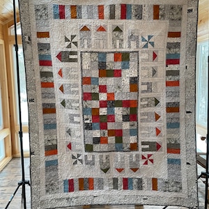 May include: A white quilt with a geometric pattern in shades of gray, brown, red, and blue. The quilt is made up of squares and rectangles, and it has a border of stripes and flowers.