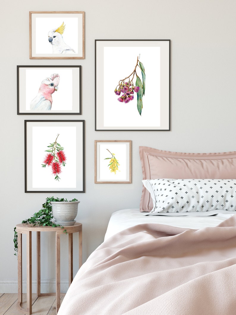 May include: A bedroom interior with a white wall and a bed with a pink blanket and pillows. There are five framed prints on the wall, featuring watercolor illustrations of Australian flora and fauna, including a pink cockatoo, a yellow cockatoo, a branch of pink flowers, a branch of red flowers, and a branch of yellow flowers.