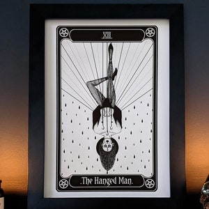 May include: Black and white illustration of The Hanged Man tarot card. The card features a figure hanging upside down with a halo and a noose around their neck. The card is framed in black and set against a dark background.