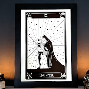 May include: Black and white illustration of The Hermit tarot card. The card features a woman in a hooded cloak holding a lantern. The card is framed in black and set against a dark background.