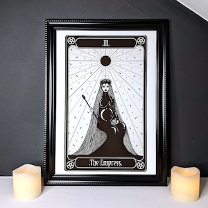 May include: A black and white illustration of The Empress tarot card. The card features a woman wearing a crown and a veil, sitting on a throne with a crescent moon and stars. The card is framed in black and set against a dark grey background.
