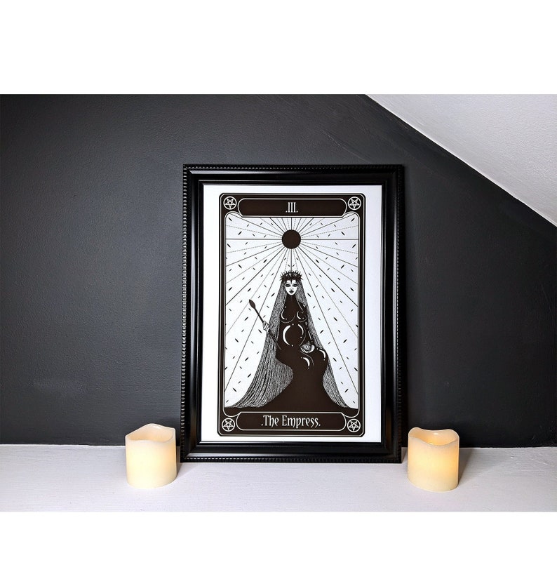 May include: A black and white illustration of The Empress tarot card. The card features a woman wearing a crown and a veil, sitting on a throne with a crescent moon and stars. The card is framed in black and set against a dark grey background.