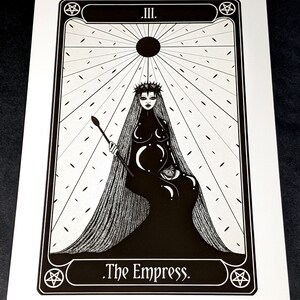 May include: Black and white illustration of The Empress tarot card. The card features a woman with a crown and long hair, sitting on a throne. The background is a radiating sun with a pentagram in the corner.
