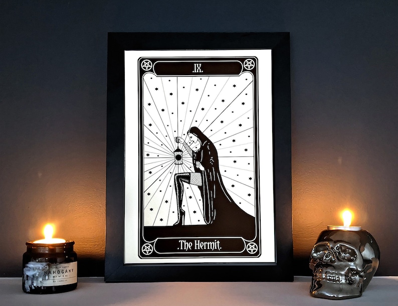 May include: Black and white illustration of The Hermit tarot card. The card features a woman in a hooded cloak holding a lantern. The card is framed in black and set against a dark background.