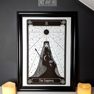 May include: Black and white illustration of The Empress tarot card. The card features a woman wearing a crown and a long flowing gown. She is holding a staff and sitting on a throne. The card is framed in black.