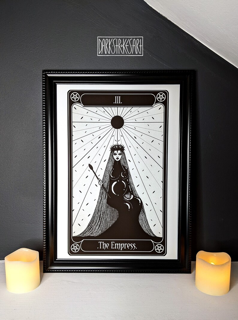 May include: Black and white illustration of The Empress tarot card. The card features a woman wearing a crown and a long flowing gown. She is holding a staff and sitting on a throne. The card is framed in black.