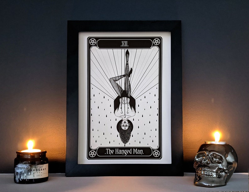 May include: Black and white illustration of The Hanged Man tarot card. The card features a figure hanging upside down with a halo and a noose around their neck. The card is framed in black and set against a dark background.